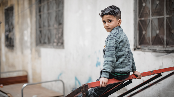 Boy sitting in front of old building looking into the camera | War Child