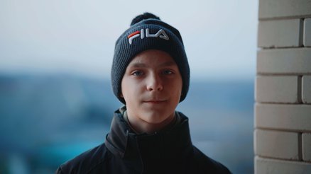 Ihor, a teenage boy from Ukraine, stands outside a flat in Moldova, slightly smiling, wearing a hat